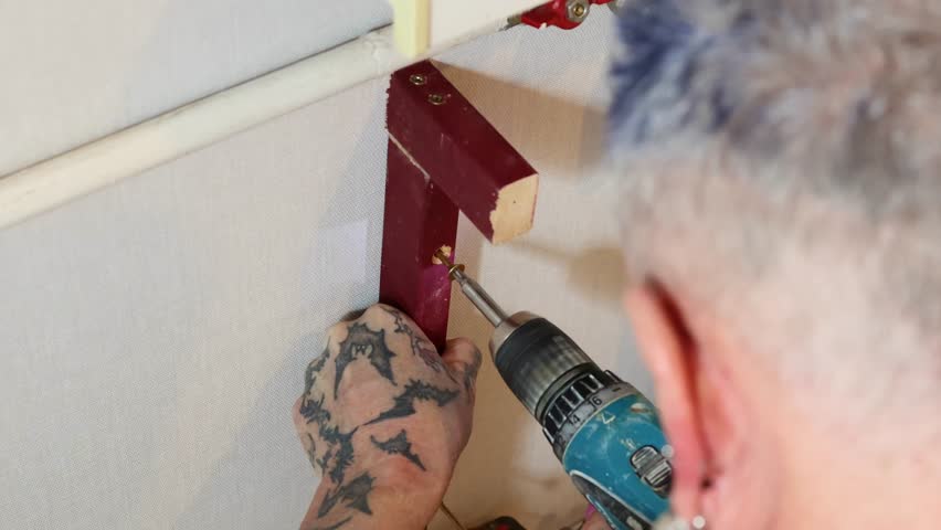 Close up of a person with hand tattoos using an electric cordless drill to drive a screw into a red wooden beam during a home renovation or DIY construction project.