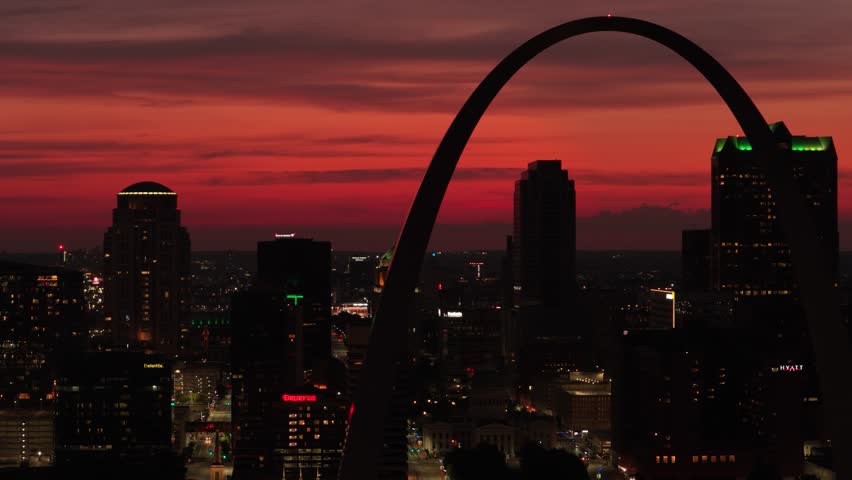 Cinematic drone footage of downtown St. Louis at sunset, with the Gateway Arch silhouetted against a bright, glowing sky as the city transitions into evening.