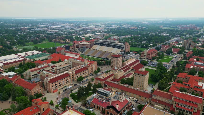 An impressive aerial shot glides over the University of Colorado Boulder campus, showcasing the distinctive red-tiled roofs of its historic buildings and the expansive Folsom Field stadium.