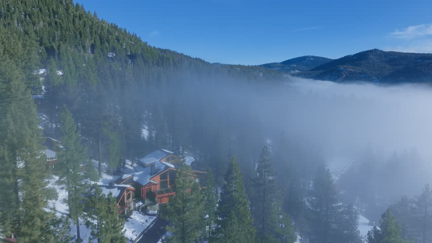 Aerial view of a mountain cabin surrounded by pine forest as low fog drifts through a winter valley at Palisades Tahoe, California, USA. Snow on the ground and clear sky create a calm alpine mood.