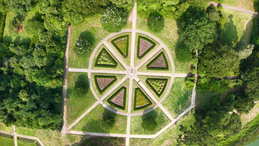 Drone aerial view of a landscaped garden in Charleston, South Carolina, featuring a circular design with radial paths dividing the lawn into wedge-shaped segments. Surrounded by trees and greenery.