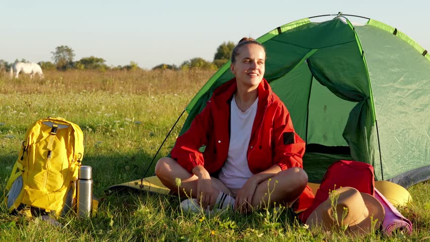 A woman sits on the grass near a green tent looking at the surroundings with a backpack and water bottle beside her on a sunny day in a field