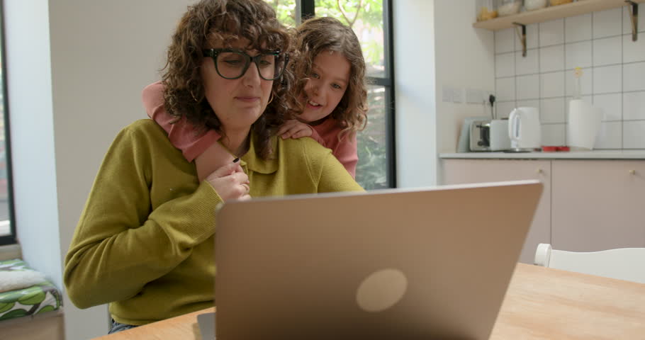 Mother at home Working on Laptop in Kitchen with Daughter Embracing