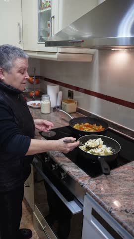 Senior woman frying vegetables in two pans on an electric stove while preparing a homemade meal