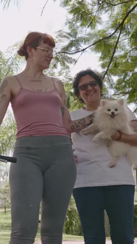 A diverse lesbian couple enjoys a leisurely walk in a lush green park on a sunny day. One woman with tattoos and red hair walks alongside her partner who is carrying a fluffy white Pomeranian dog