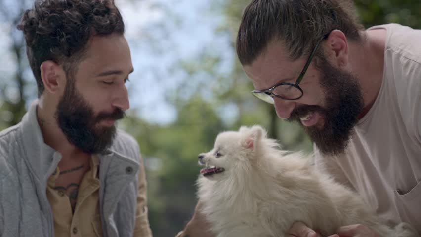 A close-up shot of two men with beards and tattoos enjoying a moment of affection with their small white dog in an outdoor setting. One man wears glasses and has his hair in a bun while they both smile warmly at their pet.