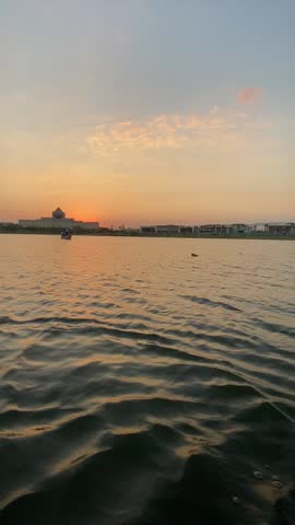 Peaceful sunset footage captured at Ain Al Sira Lake within Fustat Park, Cairo. The scene features gently rippling water reflecting warm golden and pastel sunset tones, with a small traditional boat calmly crossing the lake. In the background, the silhouette of the National Museum of Egyptian Civilization appears against the evening sky, creating a serene and cinematic urban landscape. The video conveys calmness, balance between nature and city life, and a poetic atmosphere ideal for tourism, travel, cultural, and lifestyle projects