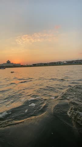 Peaceful sunset footage captured at Ain Al Sira Lake within Fustat Park, Cairo. The scene features gently rippling water reflecting warm golden and pastel sunset tones, with a small traditional boat calmly crossing the lake. In the background, the silhouette of the National Museum of Egyptian Civilization appears against the evening sky, creating a serene and cinematic urban landscape. The video conveys calmness, balance between nature and city life, and a poetic atmosphere ideal for tourism, travel, cultural, and lifestyle projects