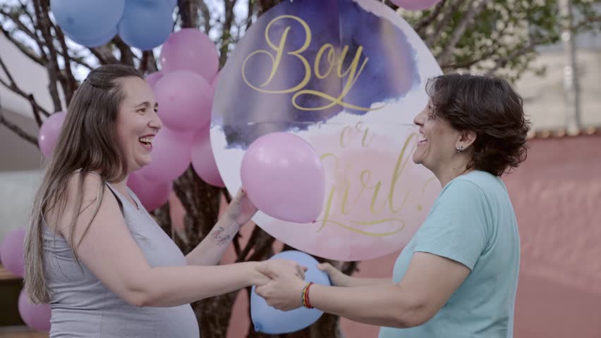 A heartwarming scene of two women sharing a joyful hug at an outdoor gender reveal party. The background is decorated with clusters of pink and blue balloons and a large sign that says 