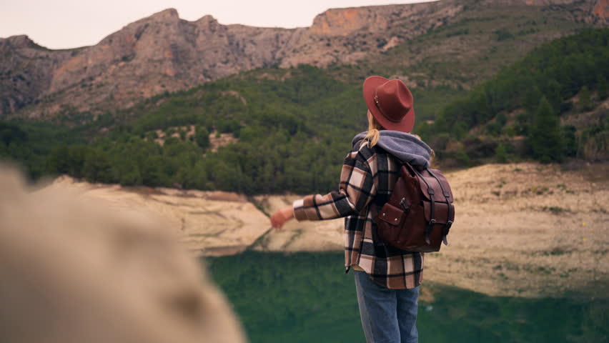 Young female hiker with backpack stand and admiring serene landscape of mountain lake, put on hat while enjoy peaceful, beautiful nature and feeling sense of freedom