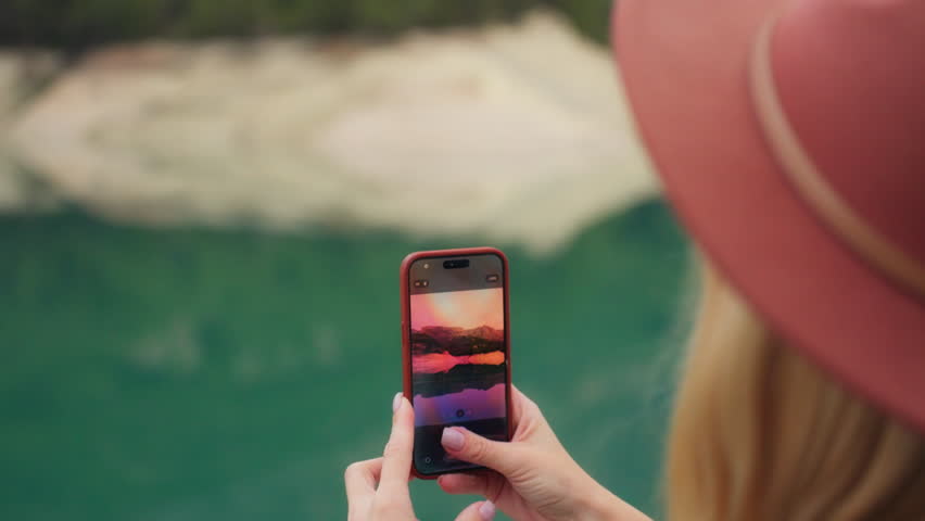 Blonde woman in  hat hold smartphone, capturing beautiful mountain landscape with turquoise lake reflecting scenery. Female traveler creating content for social media