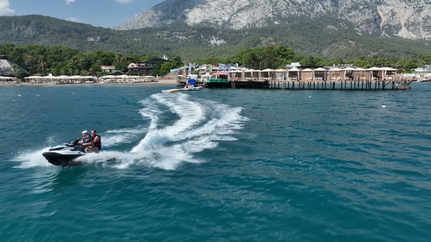A girl and a guy are riding a jet ski fast, creating big waves around them against the backdrop of a beach and high mountains. Drone shot
