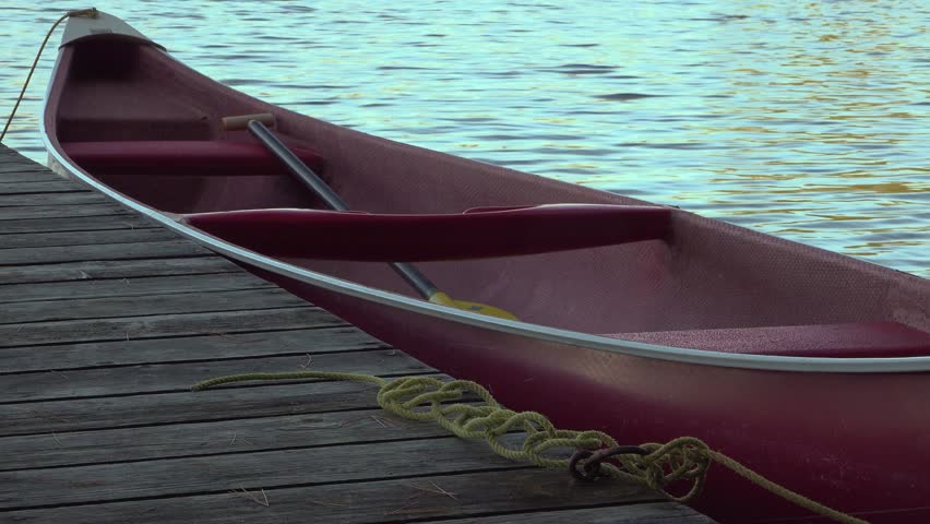 A red canoe with a paddle is tied to a weathered wooden dock as blue water glimmers beside. The peaceful lakeside scene evokes relaxation, outdoor recreation, and adventure.