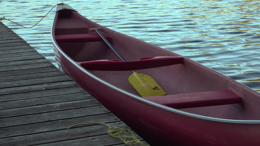 A red canoe with a paddle is tied to a weathered wooden dock as blue water glimmers beside. The peaceful lakeside scene evokes relaxation, outdoor recreation, and adventure.