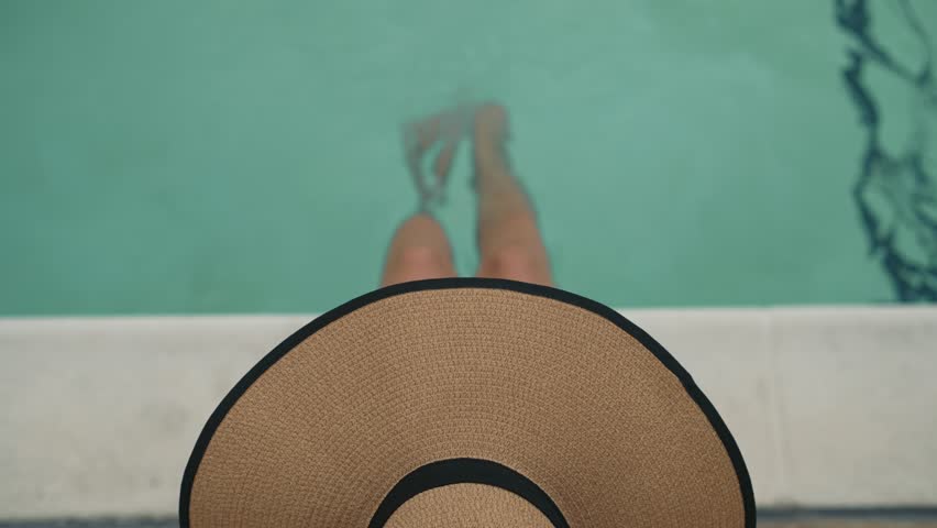 Top view of a woman in wide straw hat sitting at the edge of the pool with her legs dipped in water in a relaxed summer atmosphere