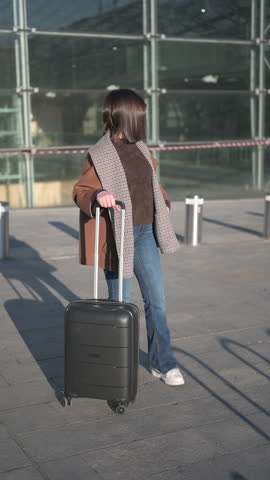 Young businesswoman with luggage standing outside a modern building, checking her phone for flight details or booking a ride-sharing service before her next business trip or vacation. Slow motion