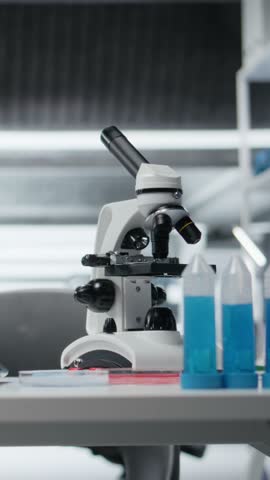 Vertical video Close up of laptop on laboratory workbench next to scientific microscope and blue liquid samples. Research station prepared for analyzing experimental data from chemical solution tests