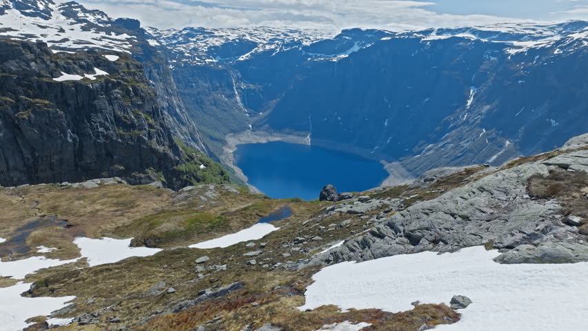  Rocky slope with snow overlooking fjord near Trolltunga, Norway. Cinematic mountain landscape with steep cliffs and deep blue water below on a clear day.
