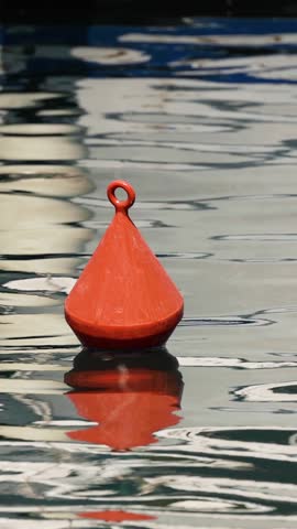 A vibrant red conical buoy bobs gently on calm, reflective water in a vertical frame.