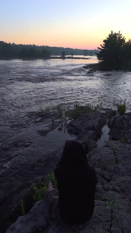 A man in a black hoodie enjoys the view while sitting on the side of a rocky riverbank surrounded by forest during a beautiful sunset.