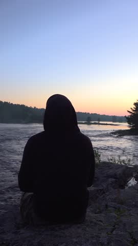 A man in a black hoodie enjoys the view while sitting on the side of a rocky riverbank surrounded by forest during a beautiful sunset.