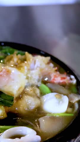 Close-up of chopsticks breaking soft egg yolk over sizzling Asian seafood noodle hot plate