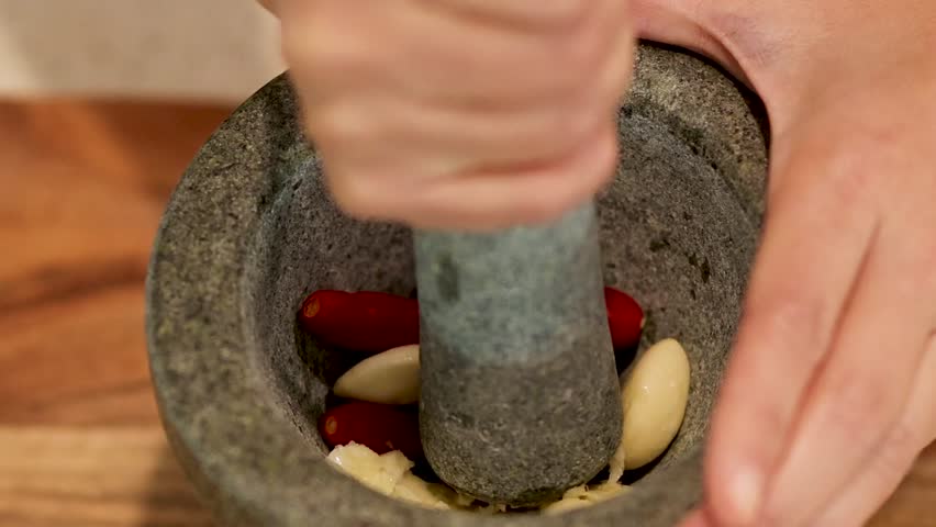A person uses a stone pestle to crush fresh garlic cloves and red chilies manually.