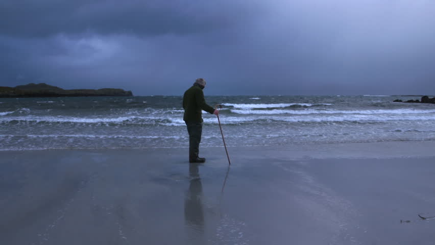 A man stands alone on the shore of a beach, gazing at the waves. The sky is dark with clouds as the sun sets. The water reflects his figure against the sand.