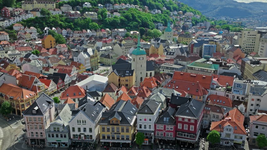 Aerial view of Bergen city center showing dense historic rooftops and colorful Nordic architecture. Prominent church tower rises above narrow streets with green hills framing the urban landscape.