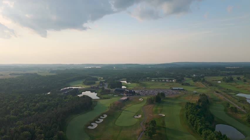 High-altitude drone panorama over a championship golf property with clubhouse, fairways, bunkers, and surrounding forest under dramatic clouds.