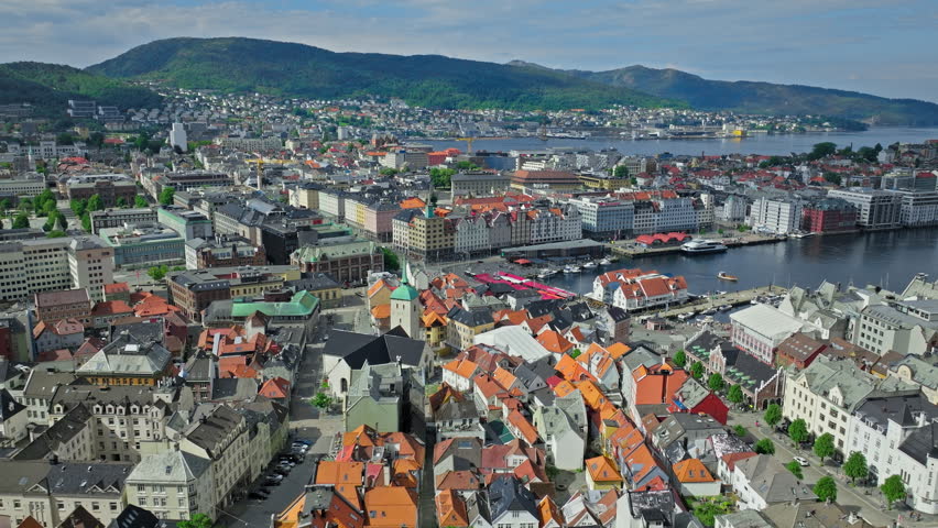 Aerial view of Bergen city center and harbor with historic rooftops in Norway. Drone footage shows dense urban architecture, colorful roofs, waterfront quays, and surrounding green hills on a clear summer day.