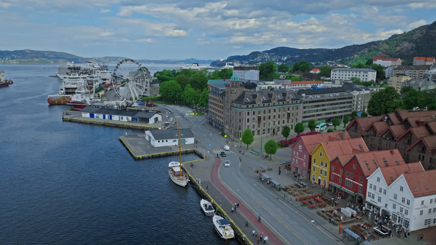 Drone view of Bergen harbor with ferries, waterfront buildings, and city infrastructure by the water in Norway. Scenic coastline and hills frame the urban landscape on a clear day.