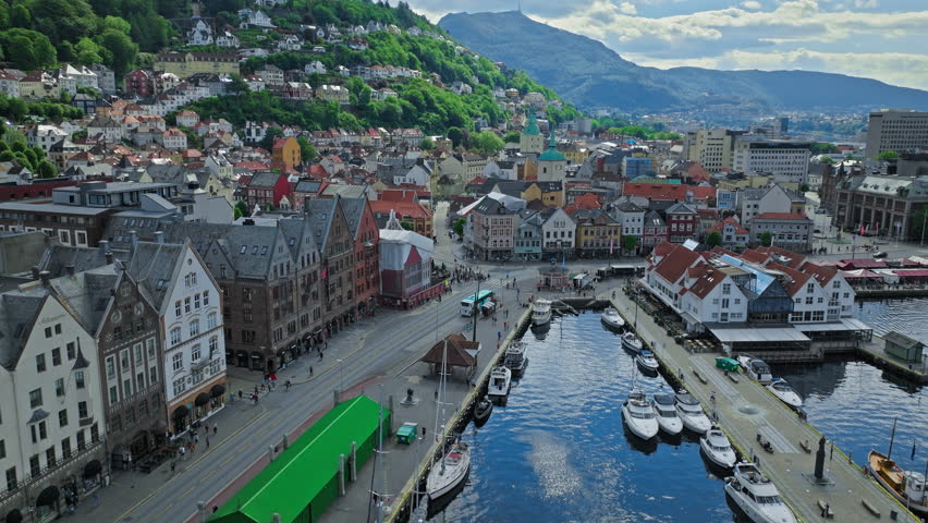 Drone aerial flying above Bergen city centre with dense rooftops and canals near the harbour in Norway. The shot captures historic urban layout surrounded by hills and fjord scenery.