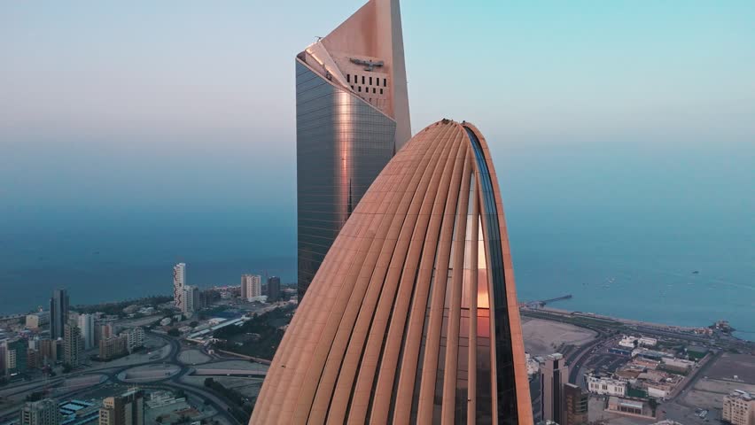 4K drone shot of modern Kuwait City seafront with iconic domed tower and glass skyscraper rising above highways and Gulf coast at blue hour, ideal for travel, corporate, and real estate projects.