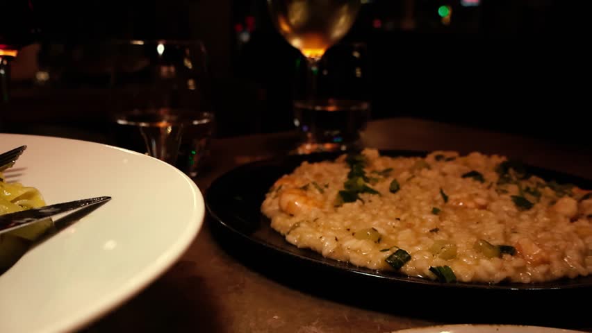 Warm restaurant scene with creamy shrimp risotto on a dark plate and wine glasses in soft bokeh light, cinematic evening dining atmosphere.