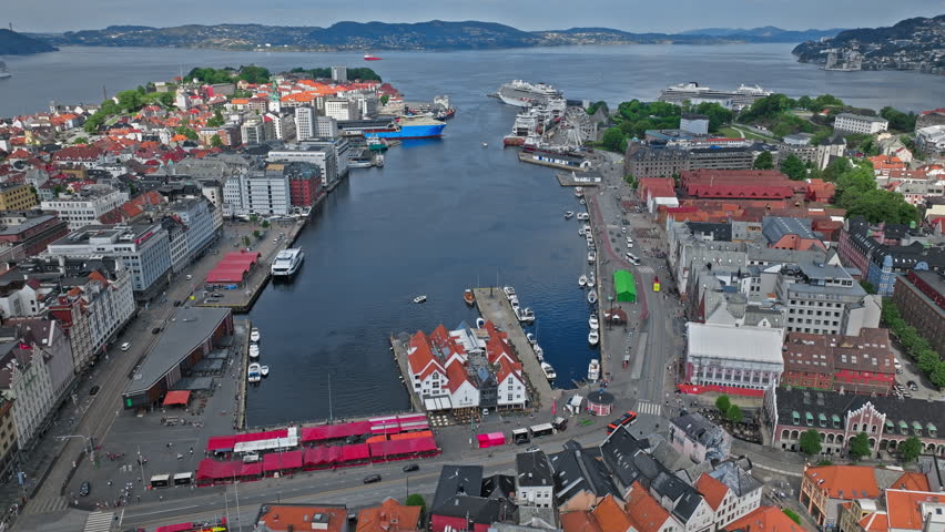 A sweeping aerial view looks down Bergen’s central harbor, where calm blue water divides the historic city, lined with colorful rooftops, quays, and busy streets. Cruise ships, ferries, and hills in the distance.