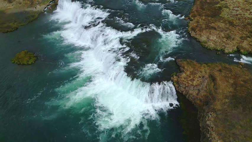 Top-down aerial shot of a large Icelandic waterfall with rushing water and foam, with green and transparent water