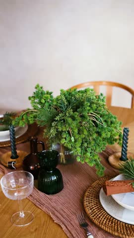 Table setting with green branches, dark vase, glass coupe, and candle on wooden table