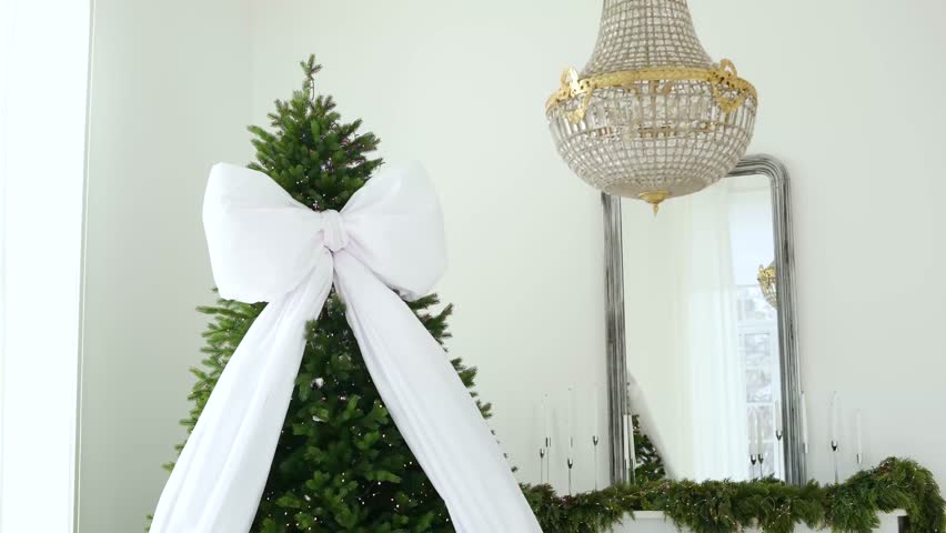 Elegant Christmas tree with large white bow, ornate chandelier, and reflective mirror background
