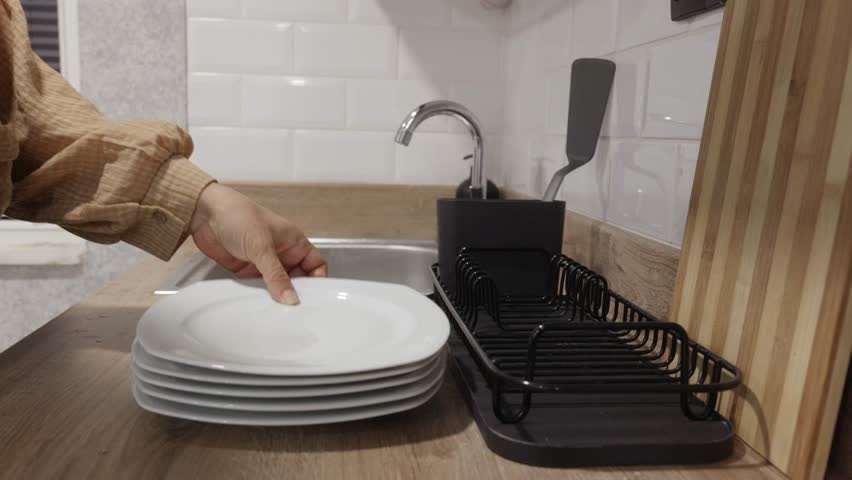 Close up view of caucasian woman hand placing clean white plate into black dish rack near kitchen sink, calm domestic atmosphere, concept of household chores.