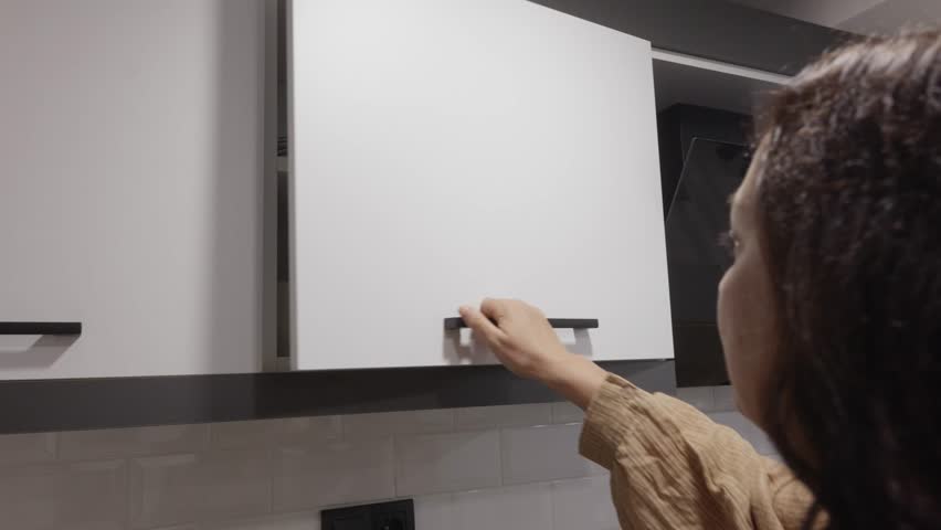 Adult caucasian woman in beige blouse standing in modern kitchen and taking white plates from cabinet, calm focused mood, symbolizing home organization and daily routine.