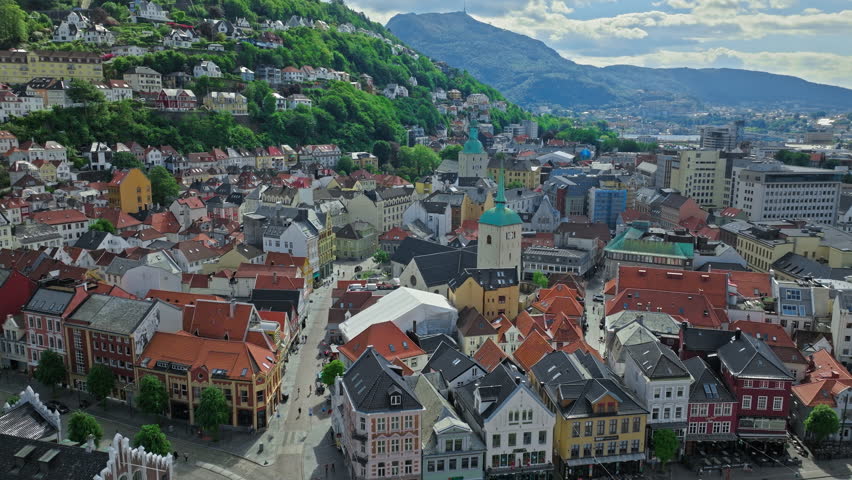 Drone aerial view over Bergen city center and harbor in Norway, showing historic waterfront buildings, boats, and busy streets. Colorful rooftops, green hills, and calm water create a lively coastal atmosphere on a clear day.