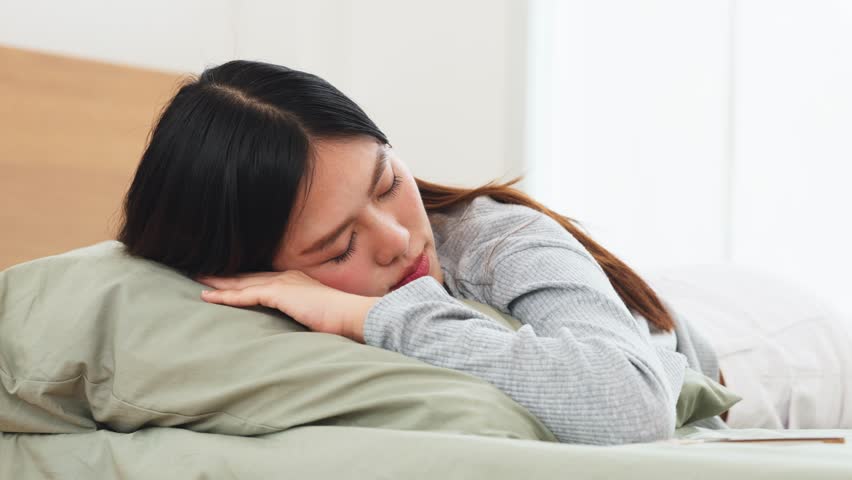 Asian young woman sleeping peacefully on pillow in bright bedroom with calm expression. Healthy sleep and wellness lifestyle concept featuring female resting during nap time or morning in domestic home setting. Perfect for bedding product advertisements, sleep disorder articles, wellness blog headers, and healthcare magazine covers. Promoting mental wellbeing, recovery, and importance of quality rest through tranquil domestic atmosphere