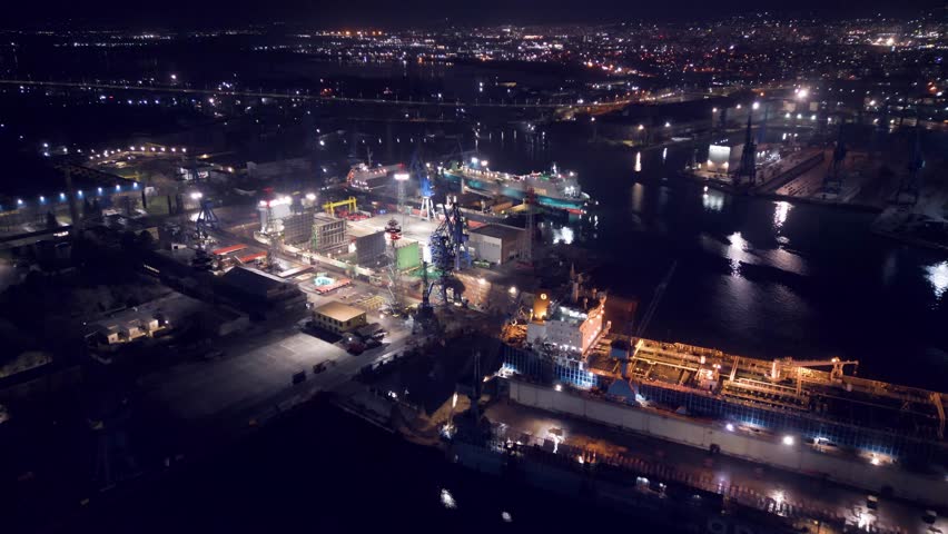 Night aerial video of a ship repair yard with dry docks, cranes and vessels illuminated by industrial lights. Large shipyard operating after dark with maritime infrastructure and harbor waters
