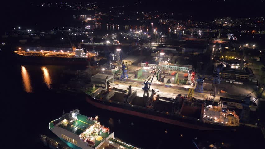 Night aerial video of a ship repair yard with dry docks, cranes and vessels illuminated by industrial lights. Large shipyard operating after dark with maritime infrastructure and harbor waters