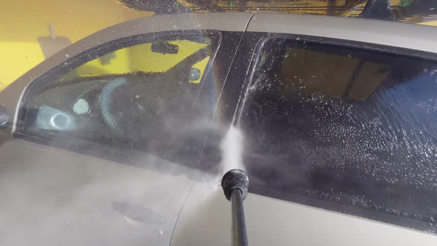 POV slow motion video of pressure washing a car side window at a self-service car wash, powerful water jet removing dirt and soap with mist and splashes