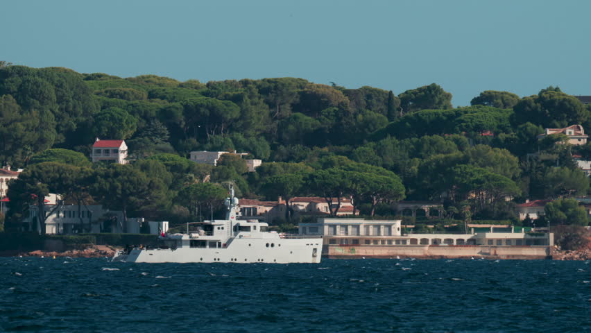 A large white vessel sails along a scenic coastline surrounded by green trees and villas