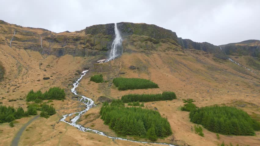 Aerial drone view of an Icelandic waterfall with a mountain backdrop on a cloudy day. Cinematic landscape showcasing untouched wilderness.