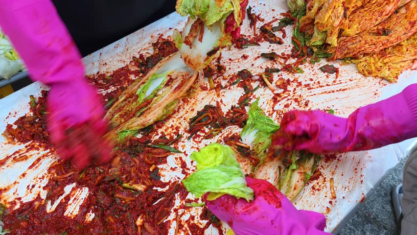 Close up of women wearing bright pink rubber gloves rubbing spicy red pepper paste onto napa cabbage leaves to make traditional kimchi on white table outdoors