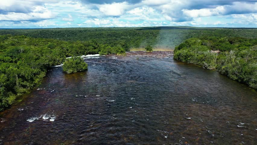 Aerial drone view of wide waterfall rapids flowing across rocky riverbed with rainbow forming above water spray surrounded by dense green forest creating vibrant natural scenery.